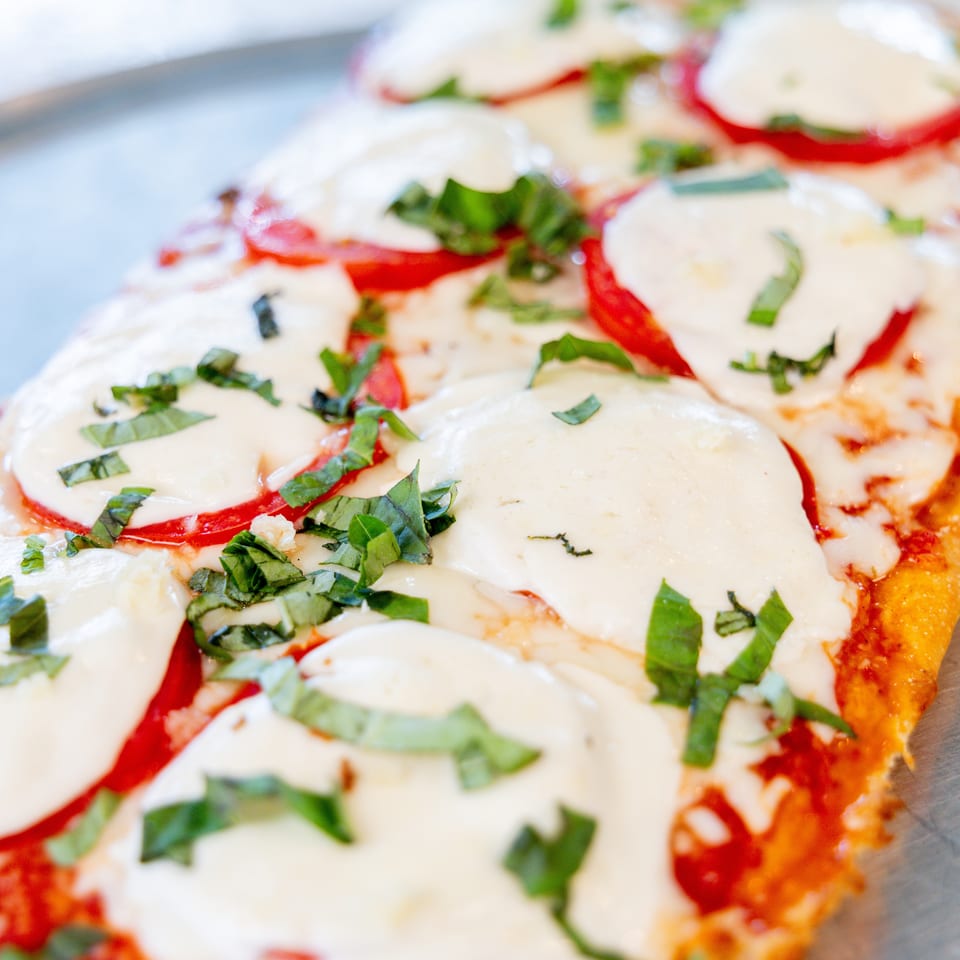 Close-up of a rectangular flatbread pizza with melted mozzarella, sliced tomatoes, and fresh basil.