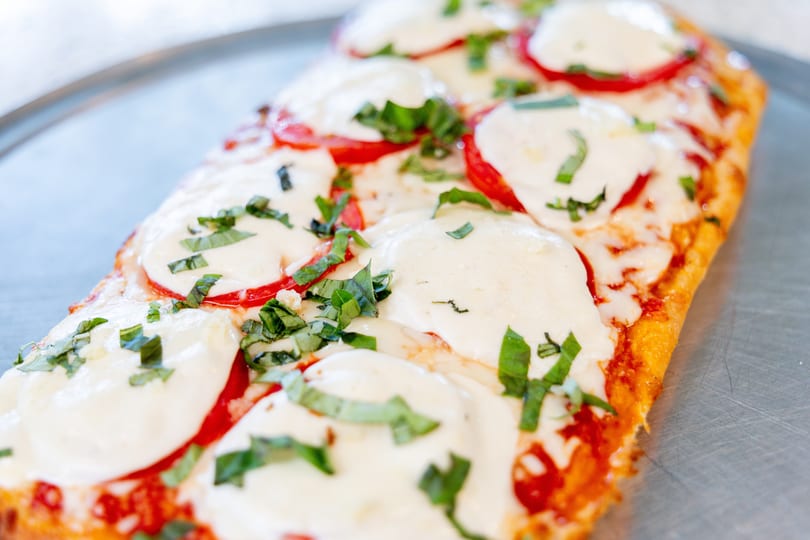 Close-up of a rectangular flatbread pizza with melted mozzarella, sliced tomatoes, and fresh basil.