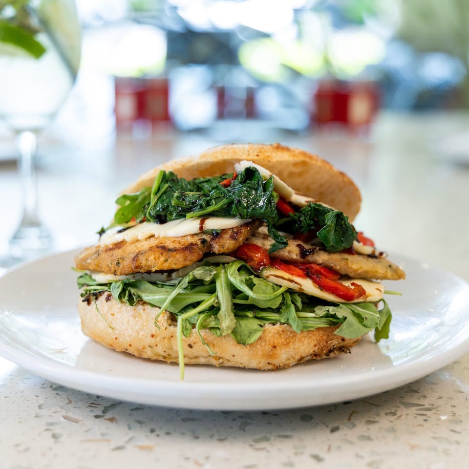 A grilled chicken sandwich with spinach, roasted red peppers, and cheese on a white plate.