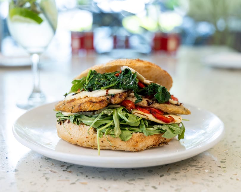 A grilled chicken sandwich with spinach, roasted red peppers, and cheese on a white plate.