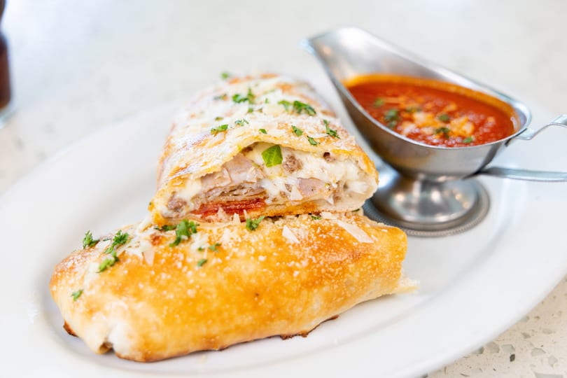 Golden-brown calzone, sliced open to reveal melted cheese, pepperoni, and sausage, served with marinara sauce.