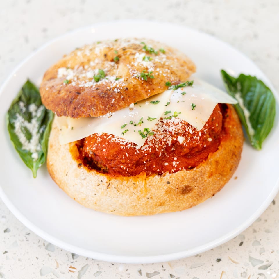 Meatball sub in a bread bowl, topped with cheese, parsley, and basil on a white plate.