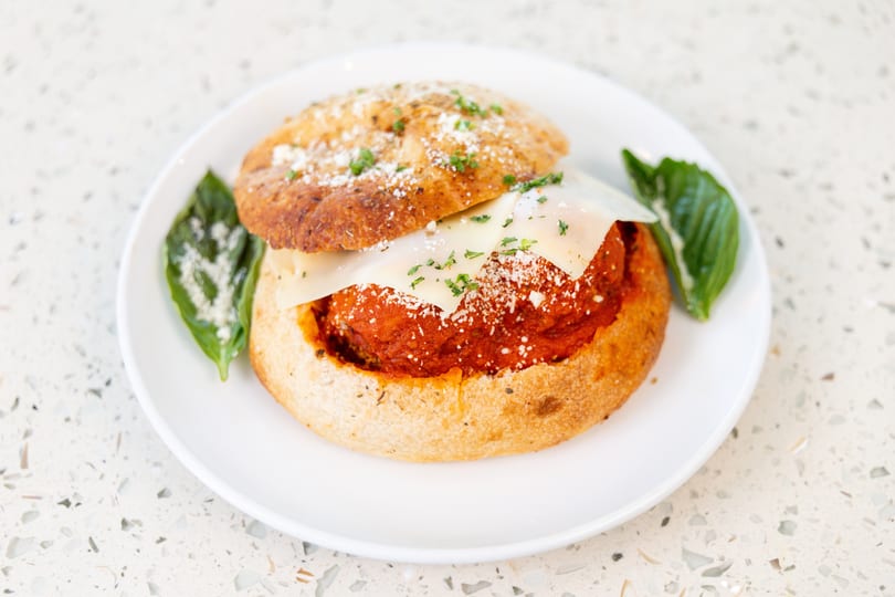 Meatball sub in a bread bowl, topped with cheese, parsley, and basil on a white plate.