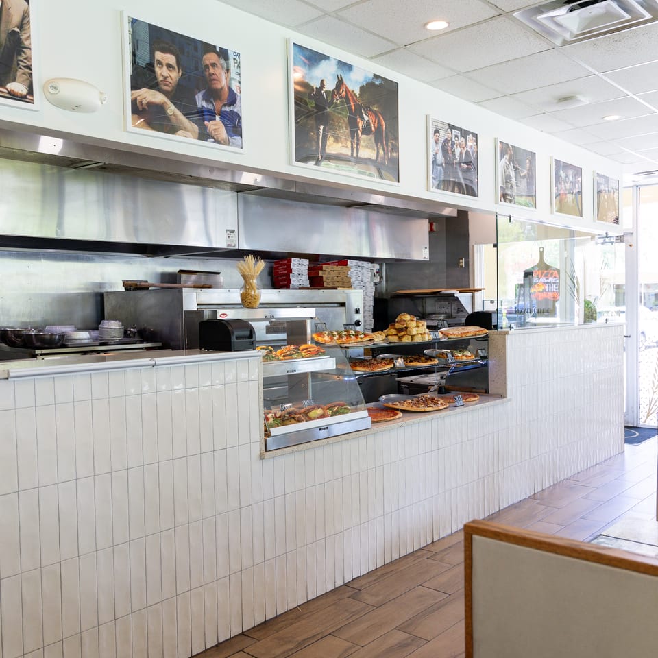 A brightly lit pizza restaurant interior with a display counter full of pizzas and baked goods.