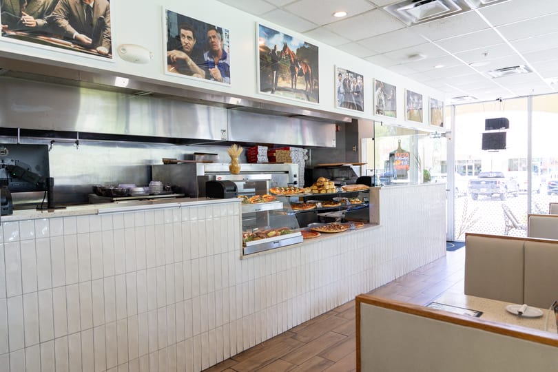 A brightly lit pizza restaurant interior with a display counter full of pizzas and baked goods.