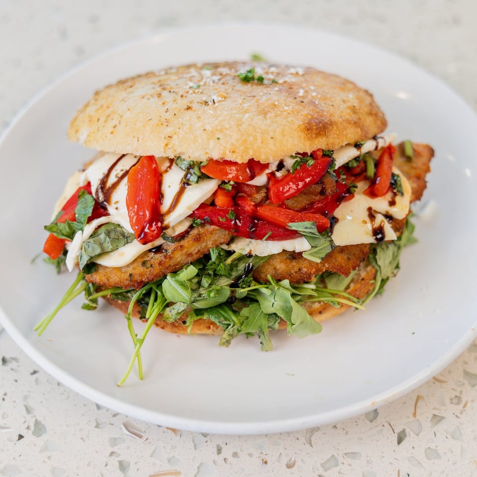 A large chicken parmesan sandwich with roasted red peppers, mozzarella, basil, and arugula on a white plate.