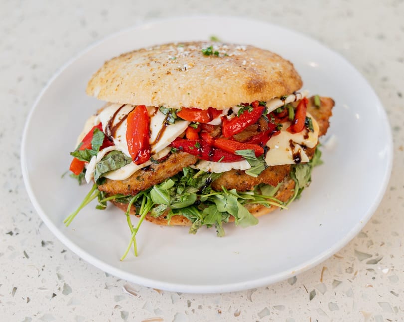 A large chicken parmesan sandwich with roasted red peppers, mozzarella, basil, and arugula on a white plate.