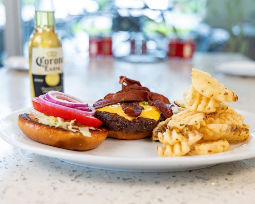 A bacon cheeseburger with waffle fries and a Corona beer bottle on a white plate.