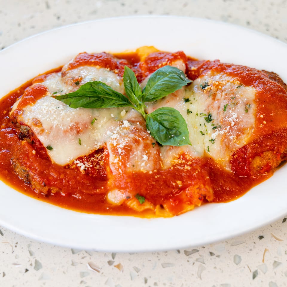 Chicken parmesan with melted mozzarella, red sauce, grated cheese, and basil on a white plate.
