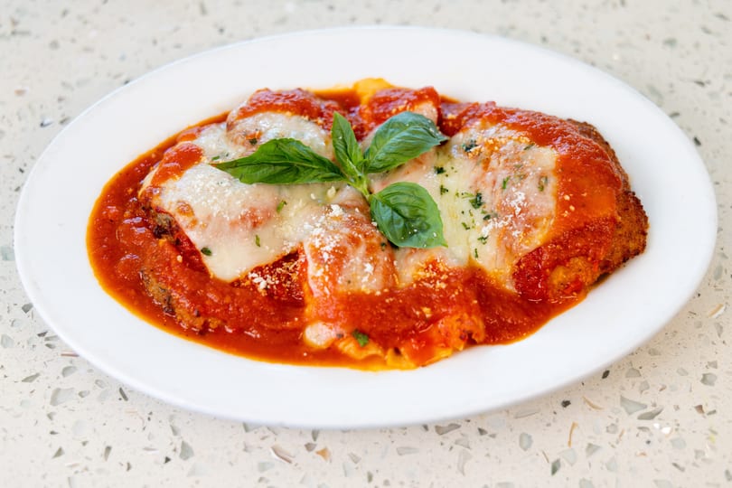Chicken parmesan with melted mozzarella, red sauce, grated cheese, and basil on a white plate.