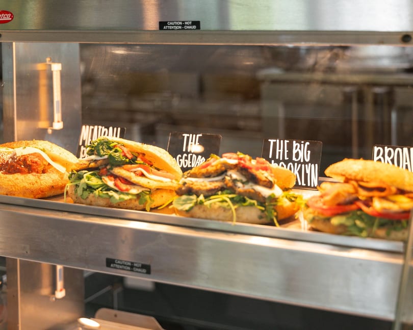 Assortment of gourmet sandwiches, including a meatball sub, displayed in a heated glass case.