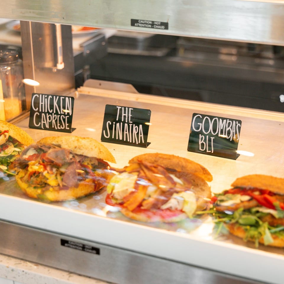 Four gourmet sandwiches with various fillings displayed in a glass deli case with name tags.