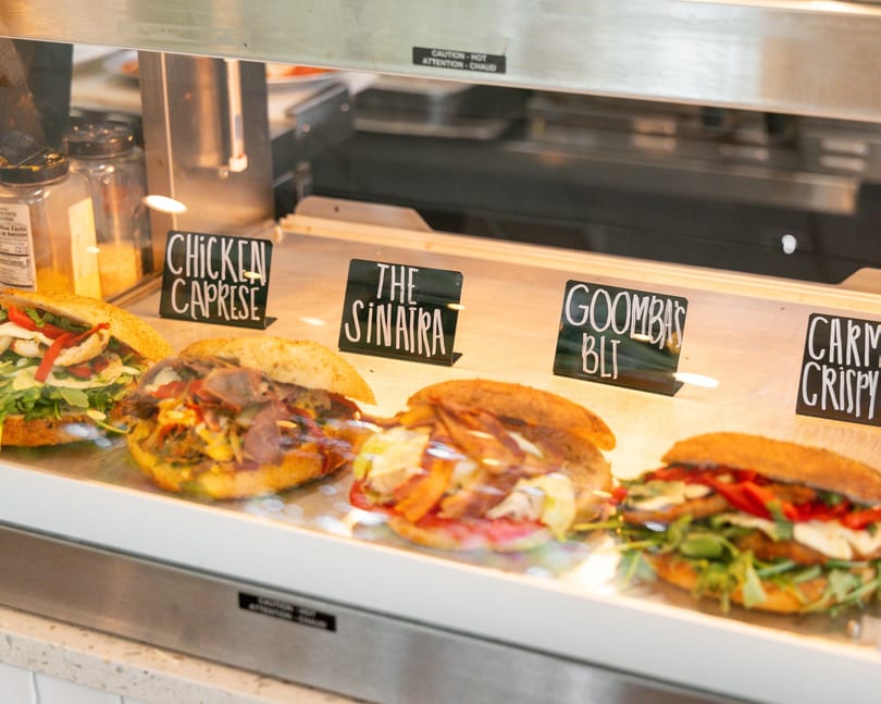 Four gourmet sandwiches with various fillings displayed in a glass deli case with name tags.