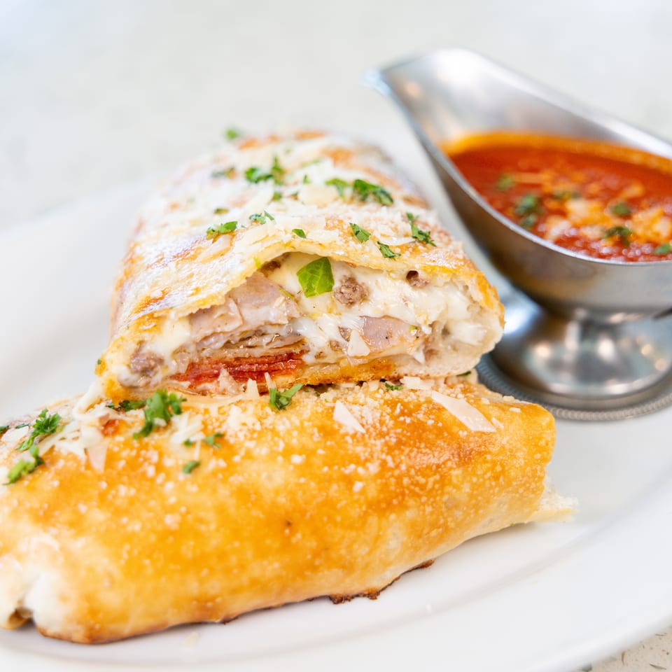 A golden-brown calzone, sliced open to reveal melted cheese and meat, served with a side of marinara sauce.