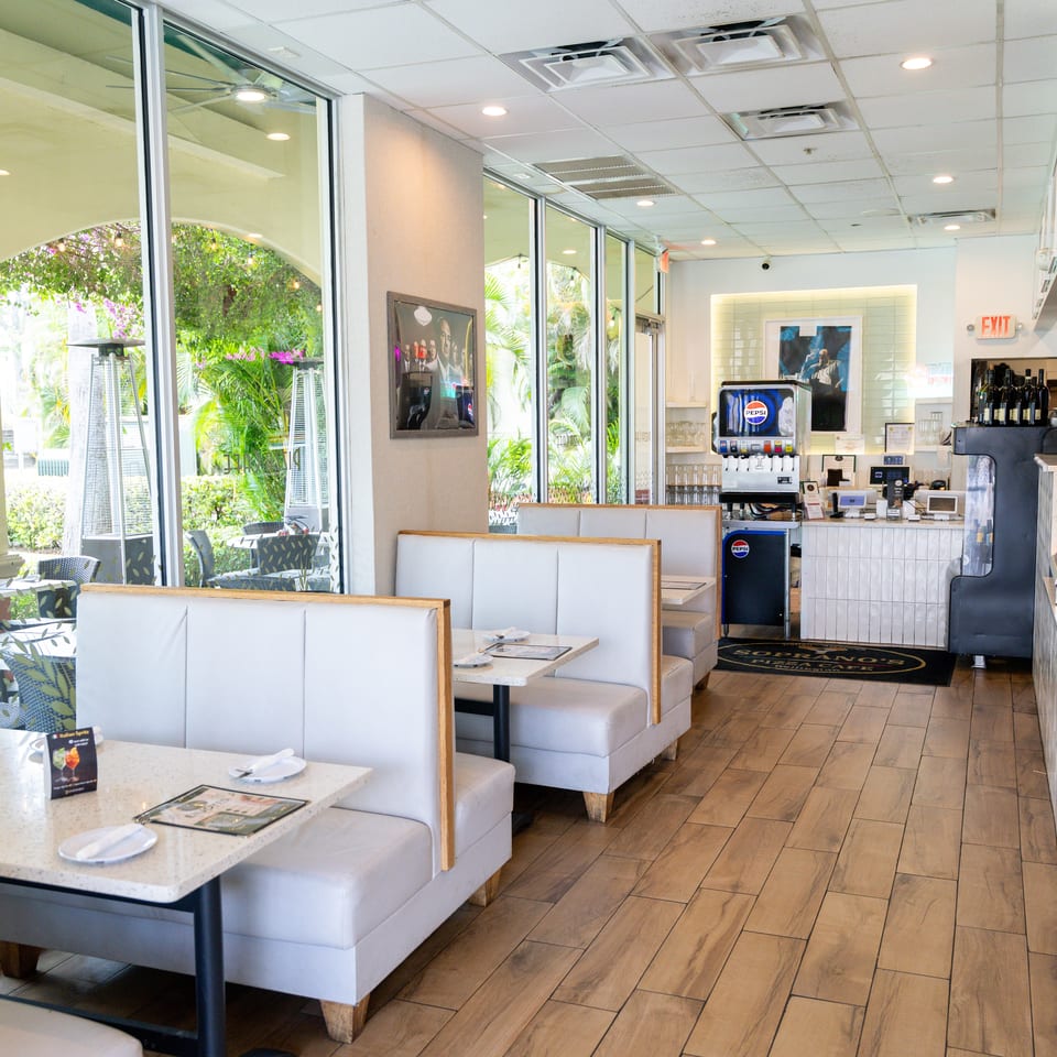 Bright, modern restaurant interior with white booths, light wood floors, and large windows overlooking outdoor seating.