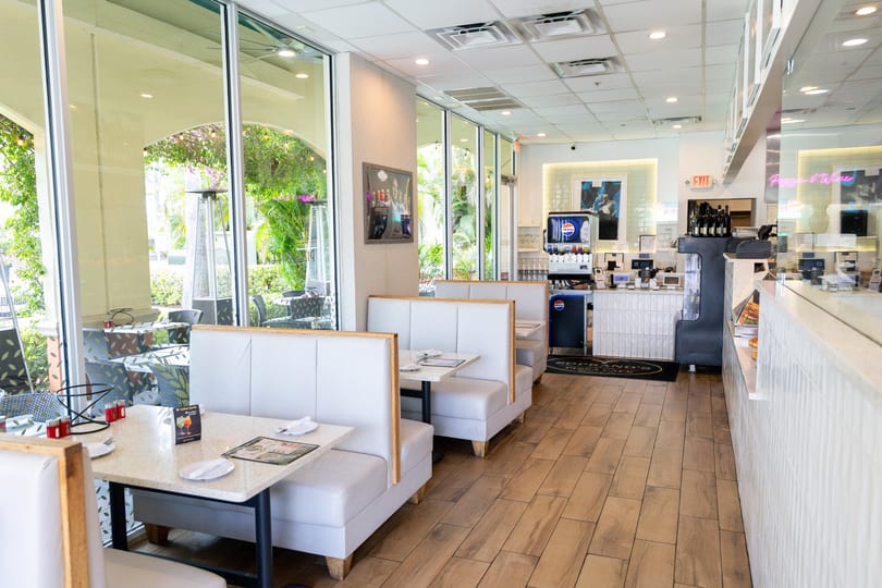 Bright, modern restaurant interior with white booths, light wood floors, and large windows overlooking outdoor seating.