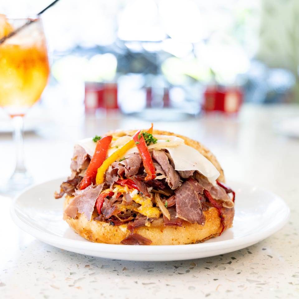 A roast beef sandwich with peppers and cheese on a white plate next to an orange cocktail.