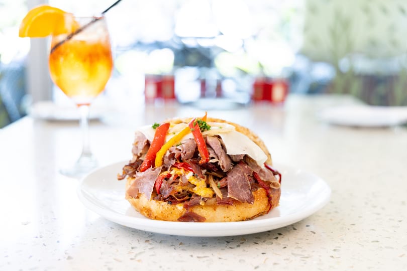 A roast beef sandwich with peppers and cheese on a white plate next to an orange cocktail.