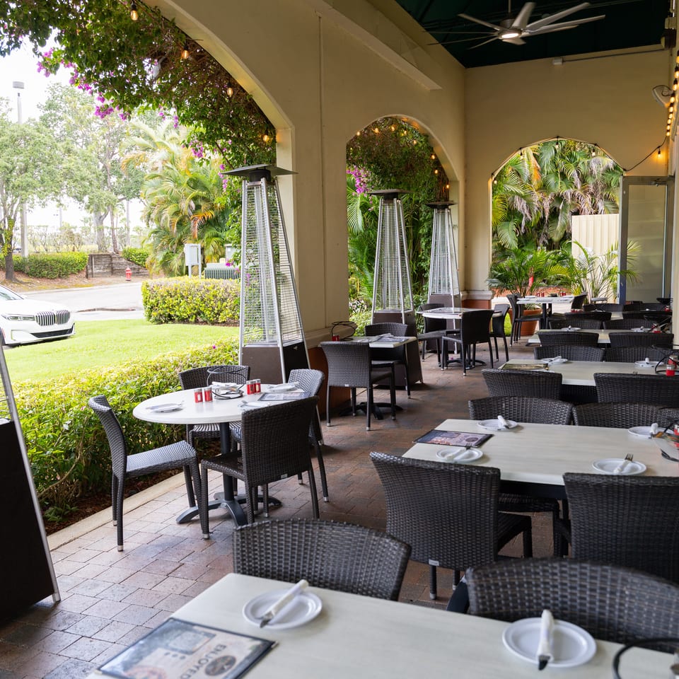An outdoor restaurant patio with tables, chairs, heaters, and lush greenery under an arched ceiling.
