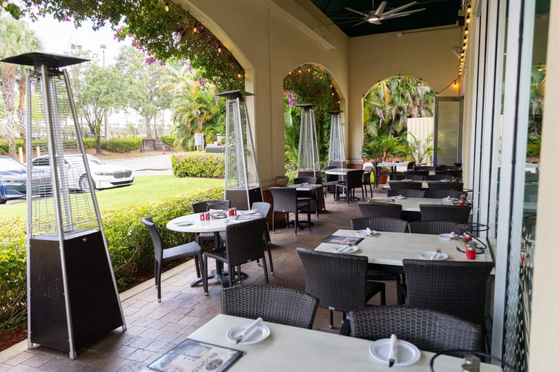 An outdoor restaurant patio with tables, chairs, heaters, and lush greenery under an arched ceiling.