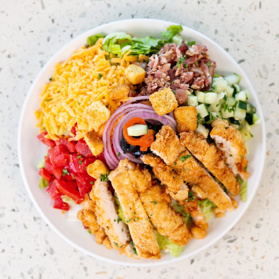 A vibrant salad with crispy chicken, bacon, cheese, tomatoes, cucumbers, and red onion on a white plate.