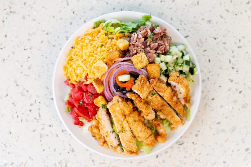 A vibrant salad with crispy chicken, bacon, cheese, tomatoes, cucumbers, and red onion on a white plate.