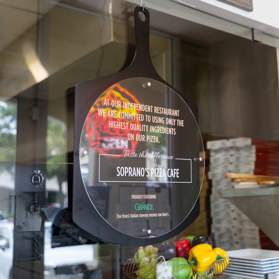 Soprano's Pizza Cafe sign in a window, featuring Grande cheese and fresh vegetables.