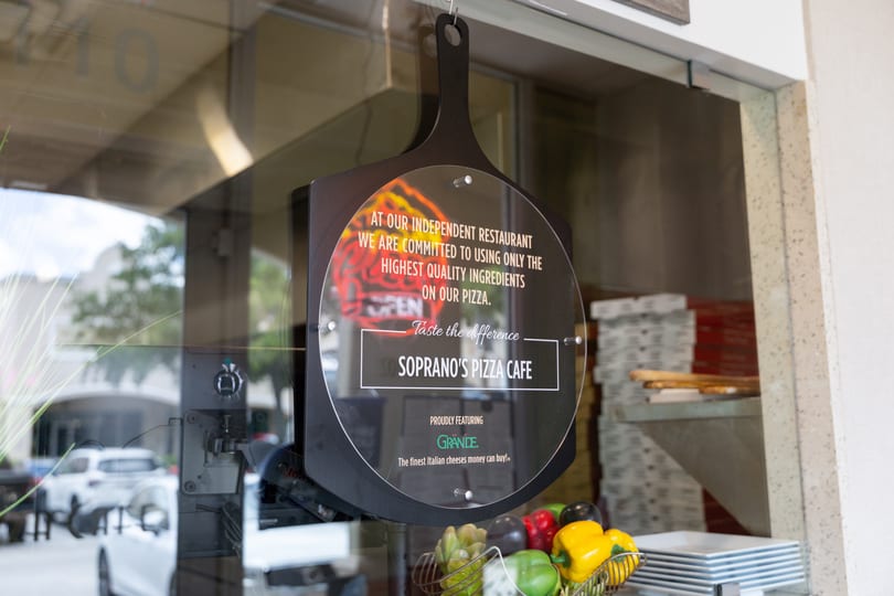 Soprano's Pizza Cafe sign in a window, featuring Grande cheese and fresh vegetables.