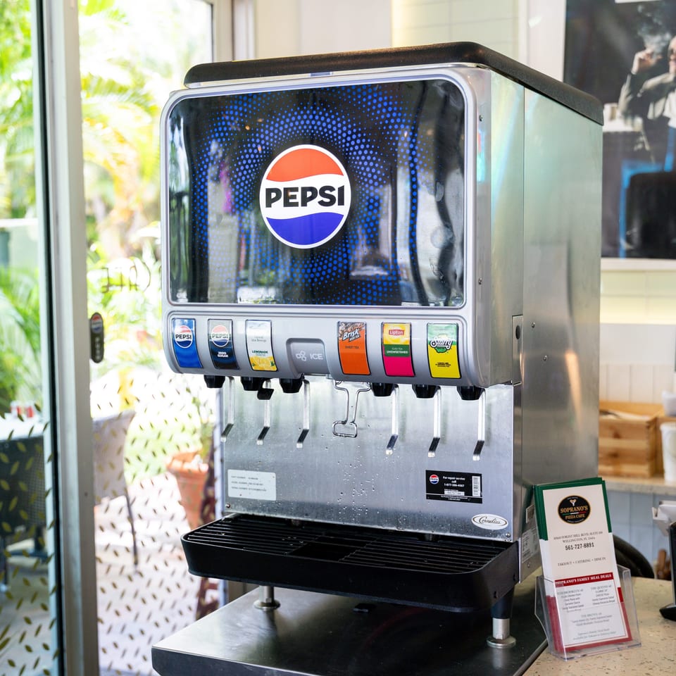 A Pepsi soda fountain with various drink options, next to a window overlooking an outdoor patio.