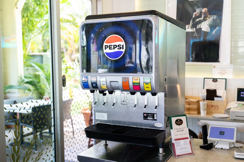 A Pepsi soda fountain with various drink options, next to a window overlooking an outdoor patio.