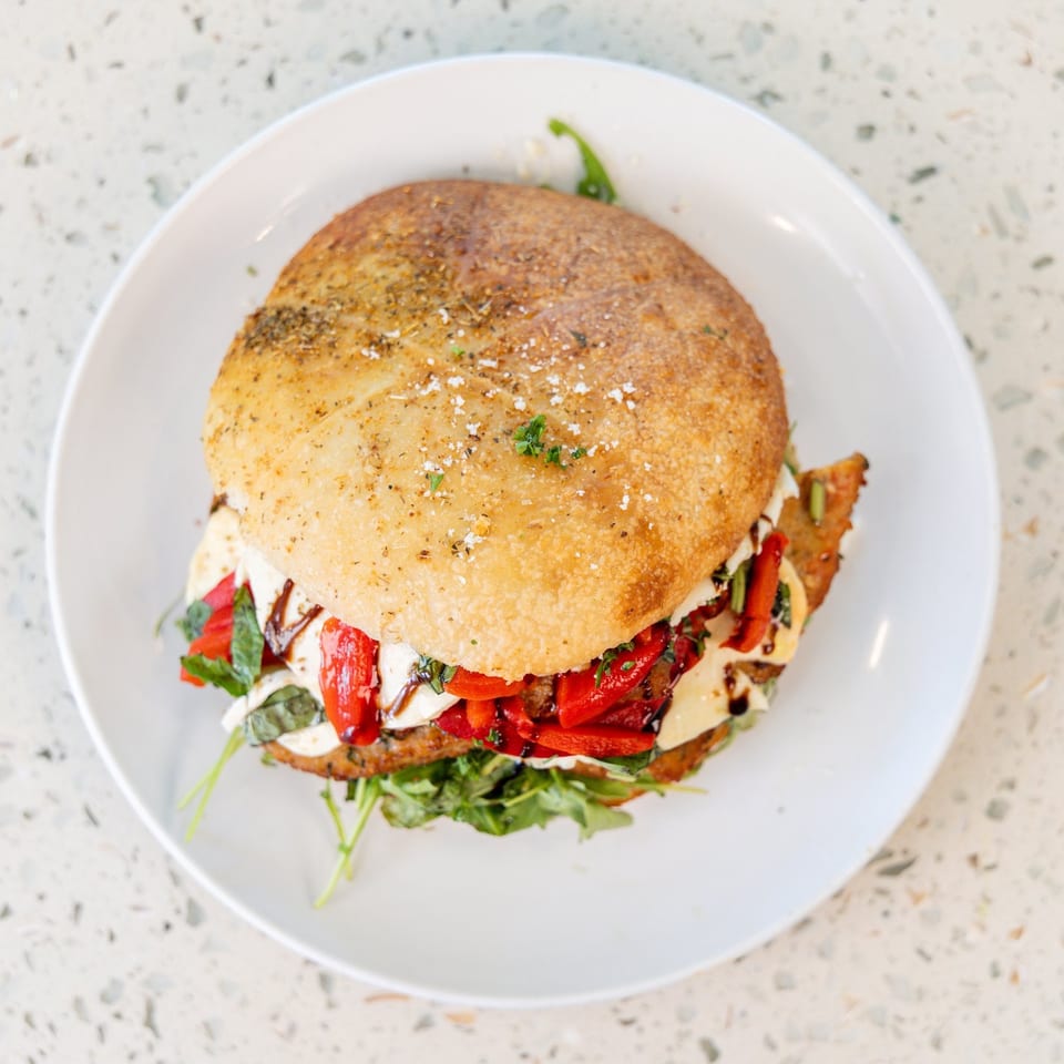A chicken parmesan sandwich with roasted red peppers, mozzarella, and balsamic glaze on a white plate.