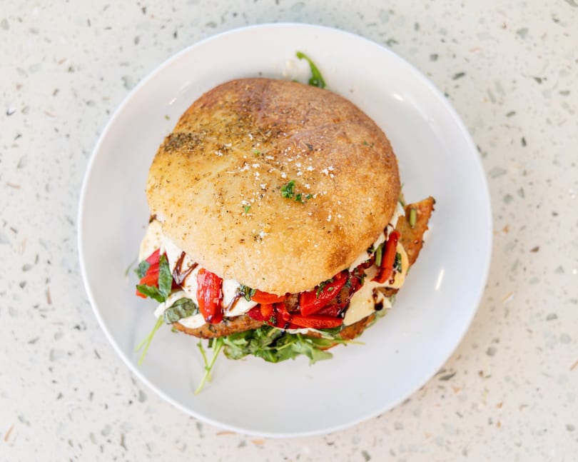 A chicken parmesan sandwich with roasted red peppers, mozzarella, and balsamic glaze on a white plate.