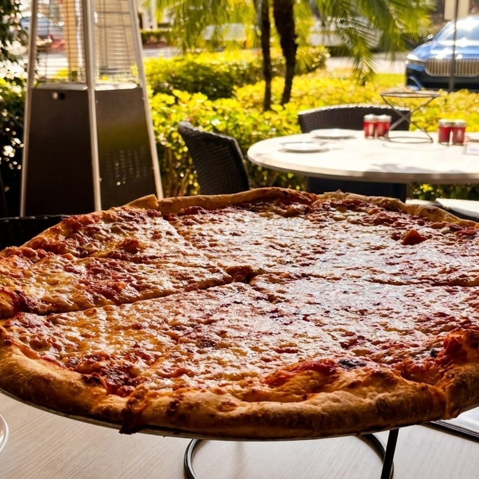 A large, sliced cheese pizza on a stand at an outdoor restaurant table with palm trees and cars in the background.