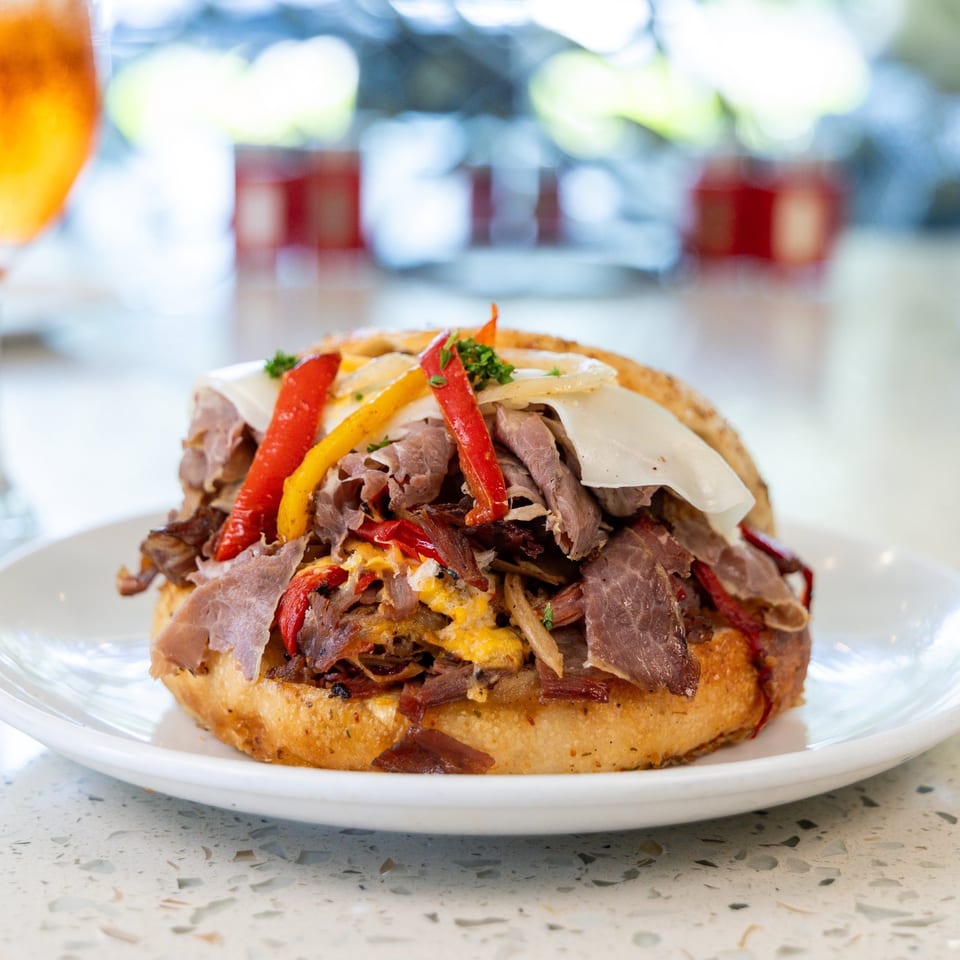 Hearty sandwich with sliced roast beef, melted cheese, colorful bell peppers, and onions on a white plate.