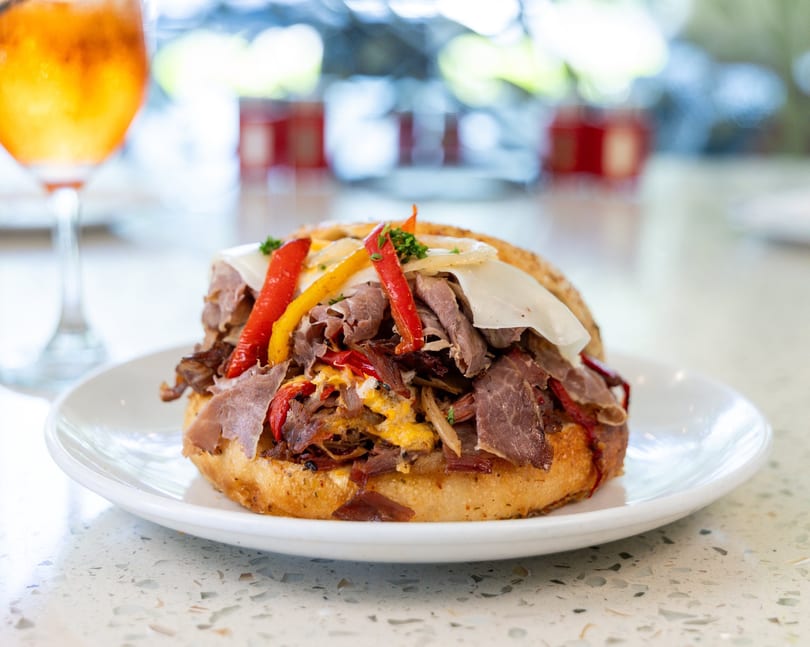 Hearty sandwich with sliced roast beef, melted cheese, colorful bell peppers, and onions on a white plate.