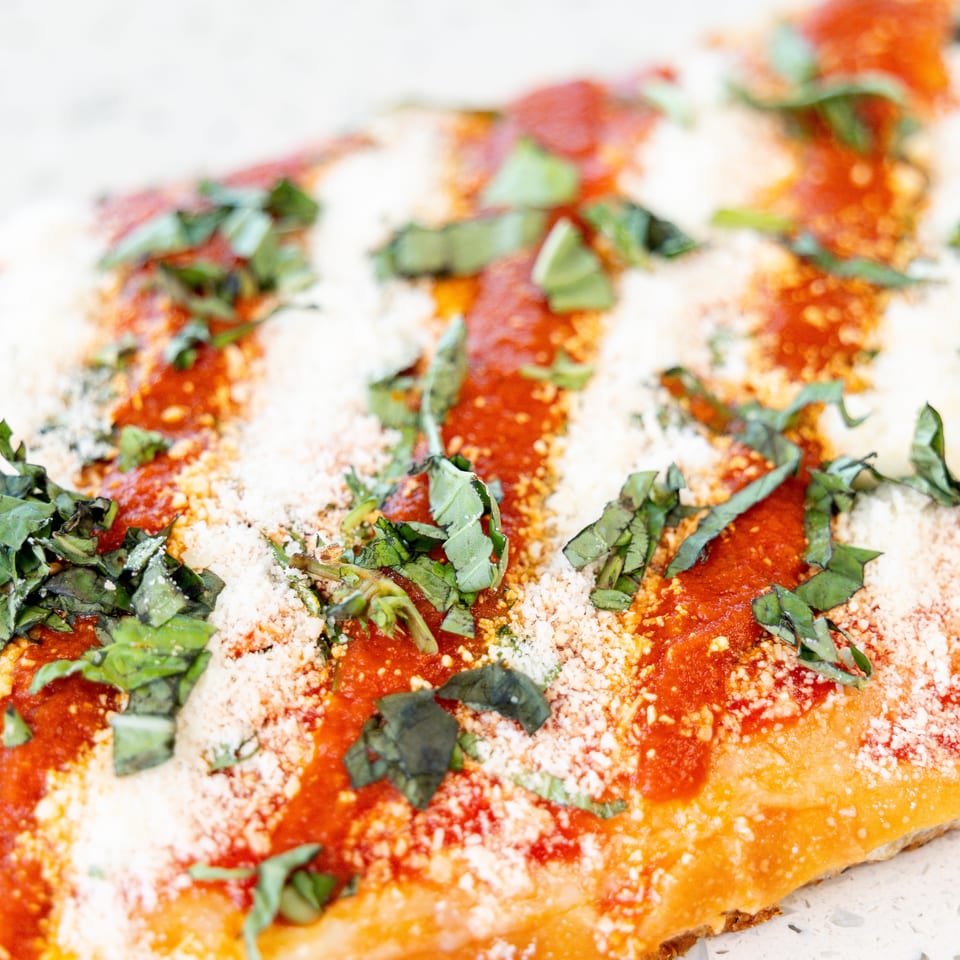 Close-up of a pizza slice with red sauce, white cheese, and green basil on a white surface.