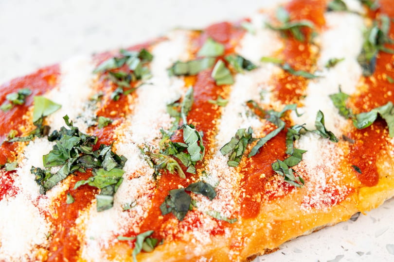 Close-up of a pizza slice with red sauce, white cheese, and green basil on a white surface.